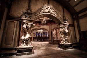 Ornate stone archway with detailed carvings and decorative statues leads to a dimly lit dining area in a themed restaurant.