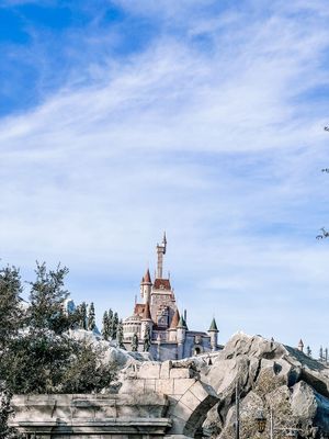 Fairy tale castle with spires and turrets sits atop rocky outcrops against a bright blue sky with wispy clouds at Disney park.