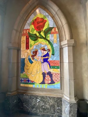 Four stained glass church windows depicting scenes from Beauty and the Beast in ornate stone archways.