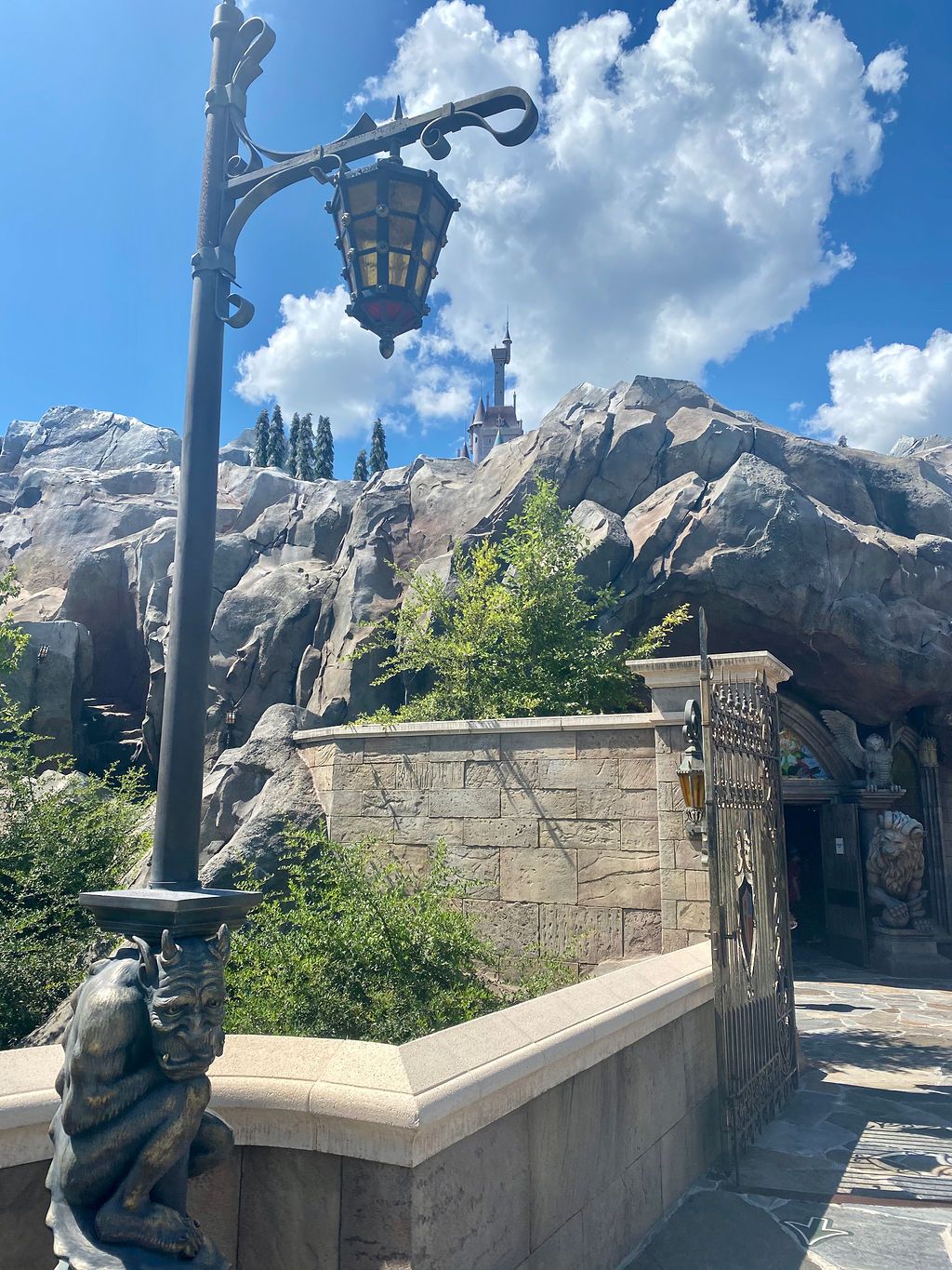 Ornate lamp post and stone wall in front of rocky mountain peaks at Disney World's Be Our Guest attraction on sunny day.