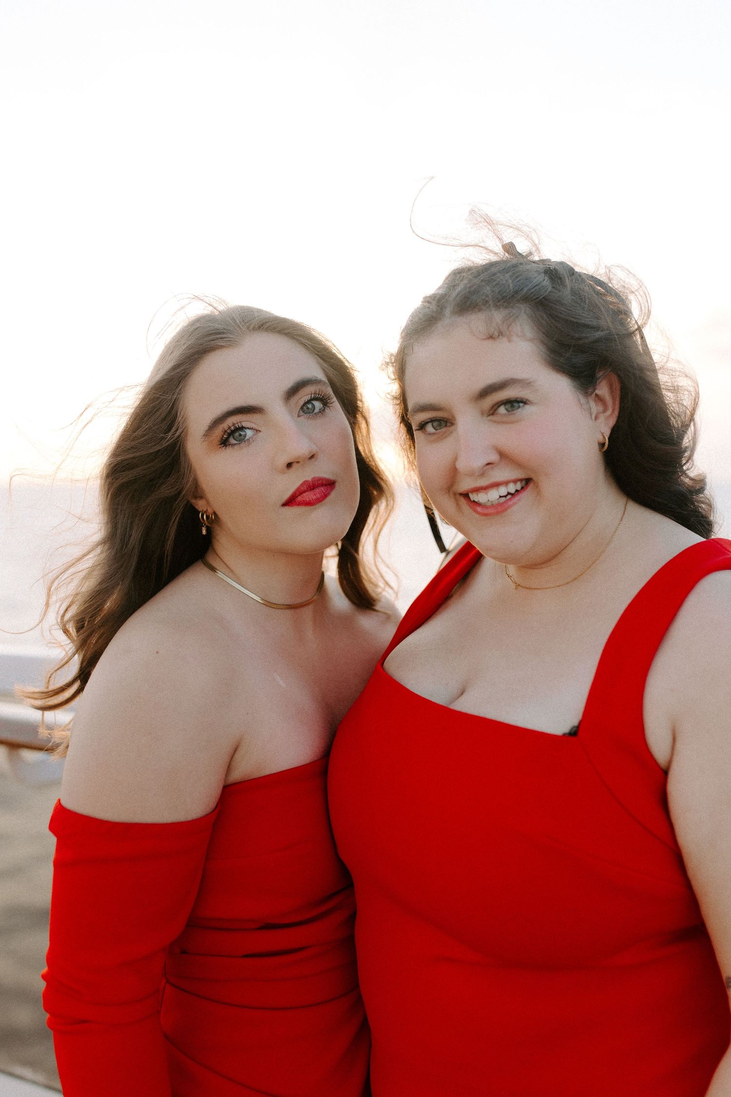 Two people in matching red off-shoulder dresses pose together at sunset.