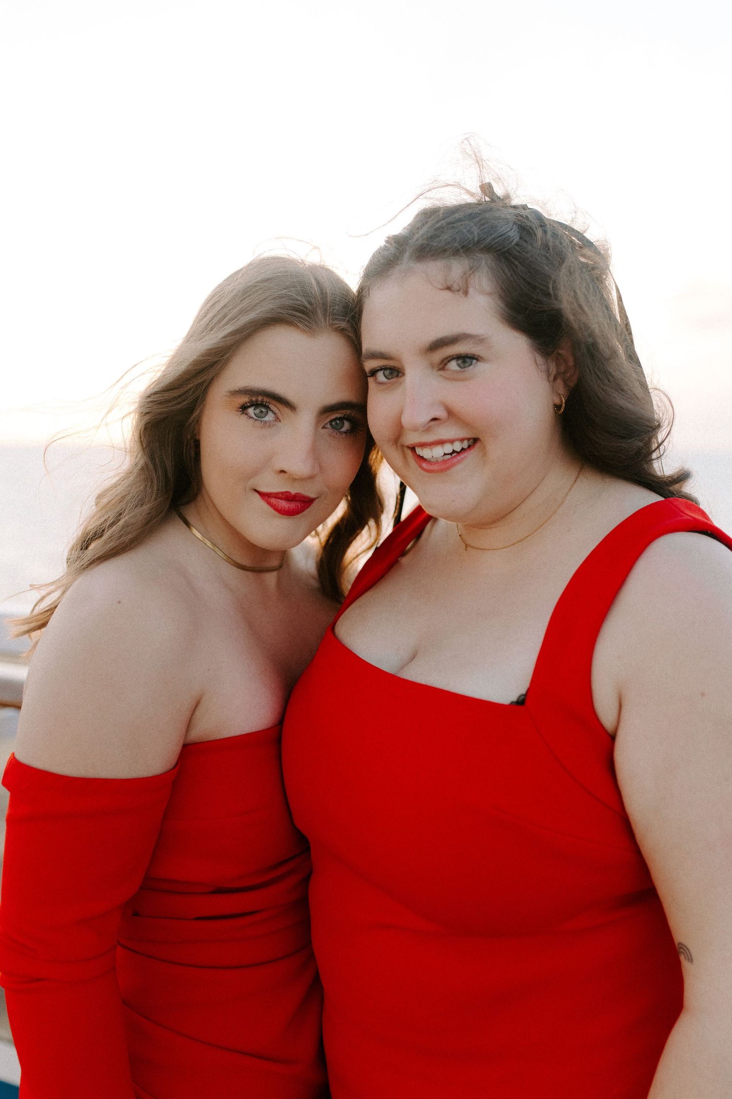 Two friends in matching red dresses share a joyful moment together against a bright background.