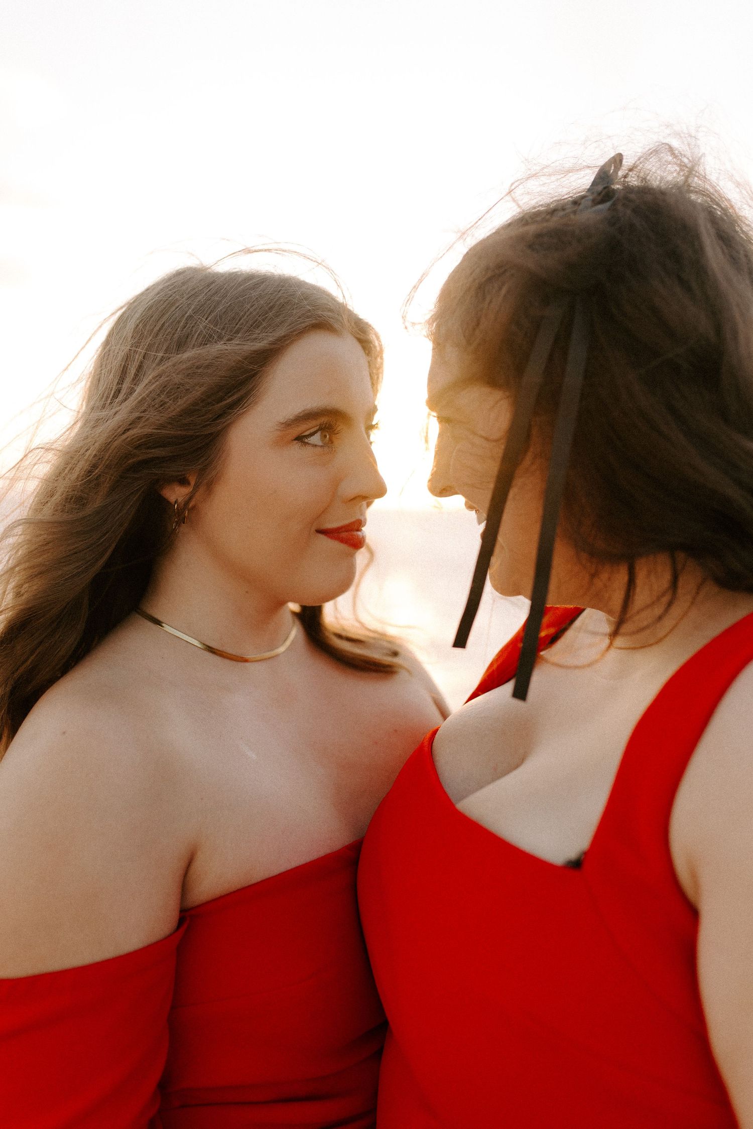 Two people in red dresses share an intimate moment as sunlight streams behind them.