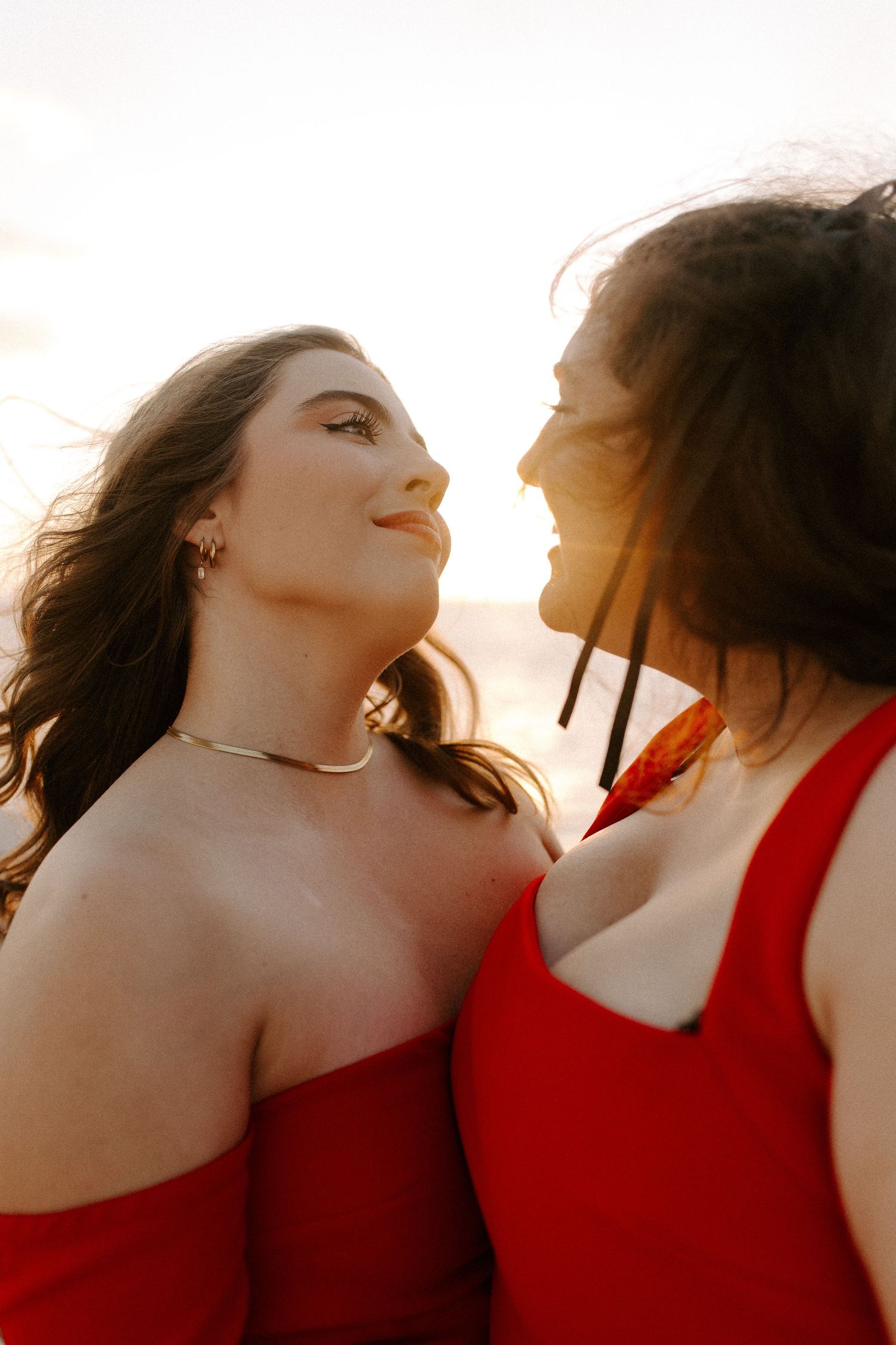 Candid portrait of two people in red dresses laughing together in warm sunlight.