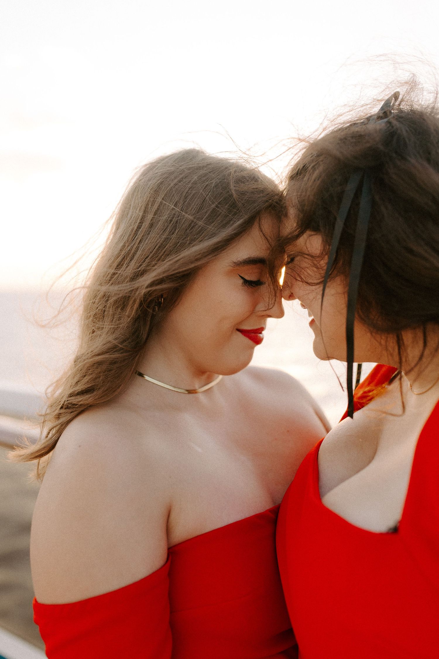 Tender portrait of two people in red dresses with faces close together at sunset.