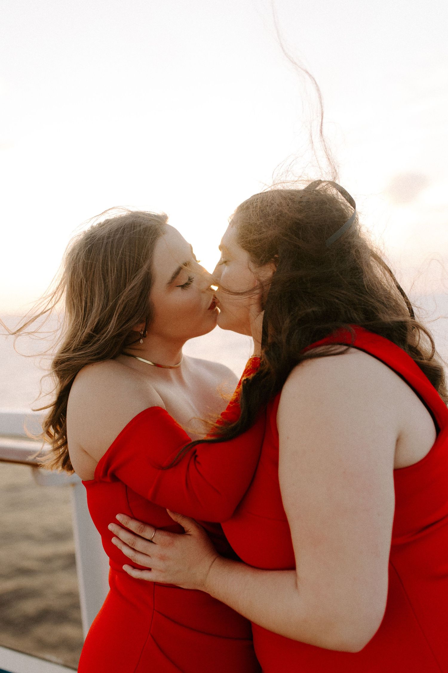 A loving couple dressed in red embracing and kissing on a cruise ship deck at sunset.