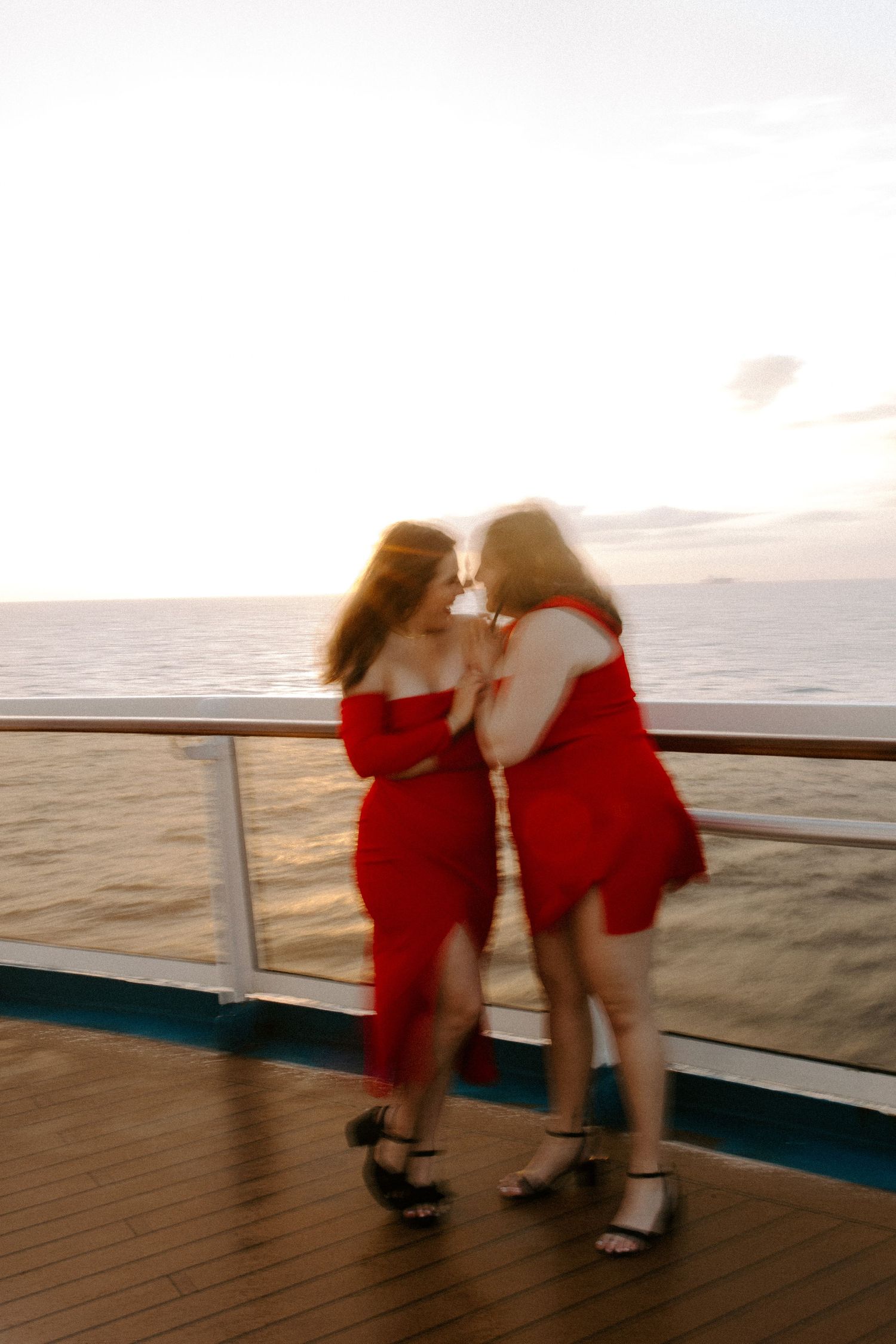Two friends in matching red dresses share a tender moment at sunset on a cruise ship deck.
