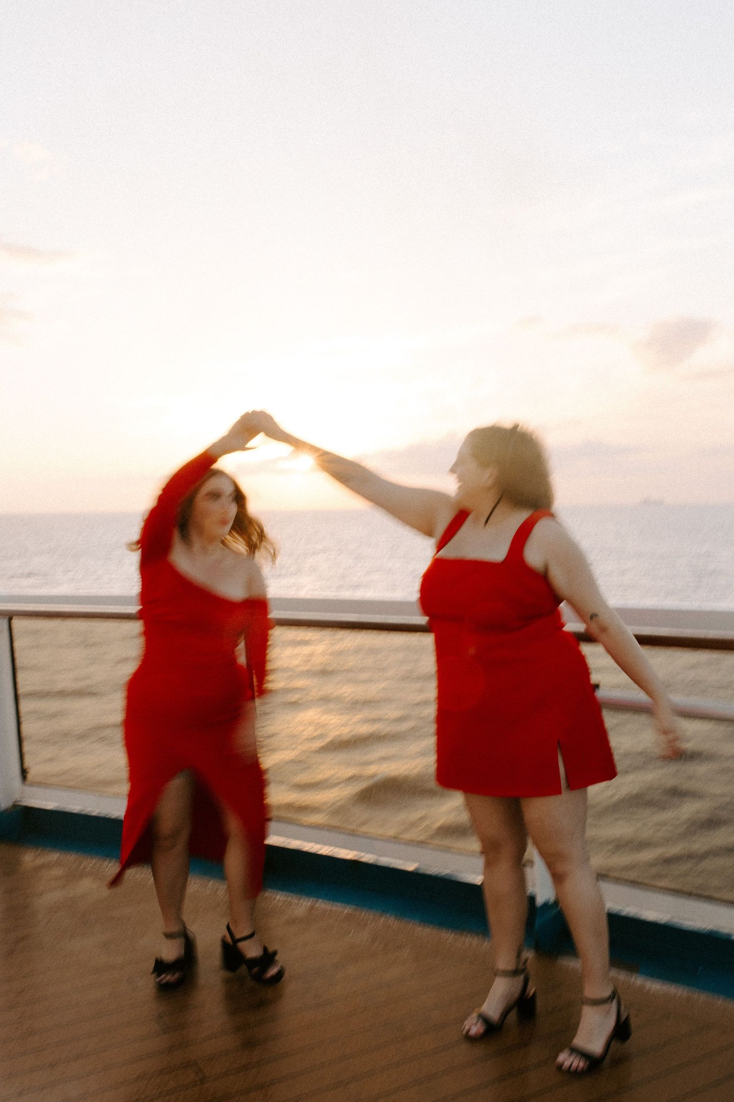 Friends in red dresses dancing and twirling on a cruise ship deck at sunset.