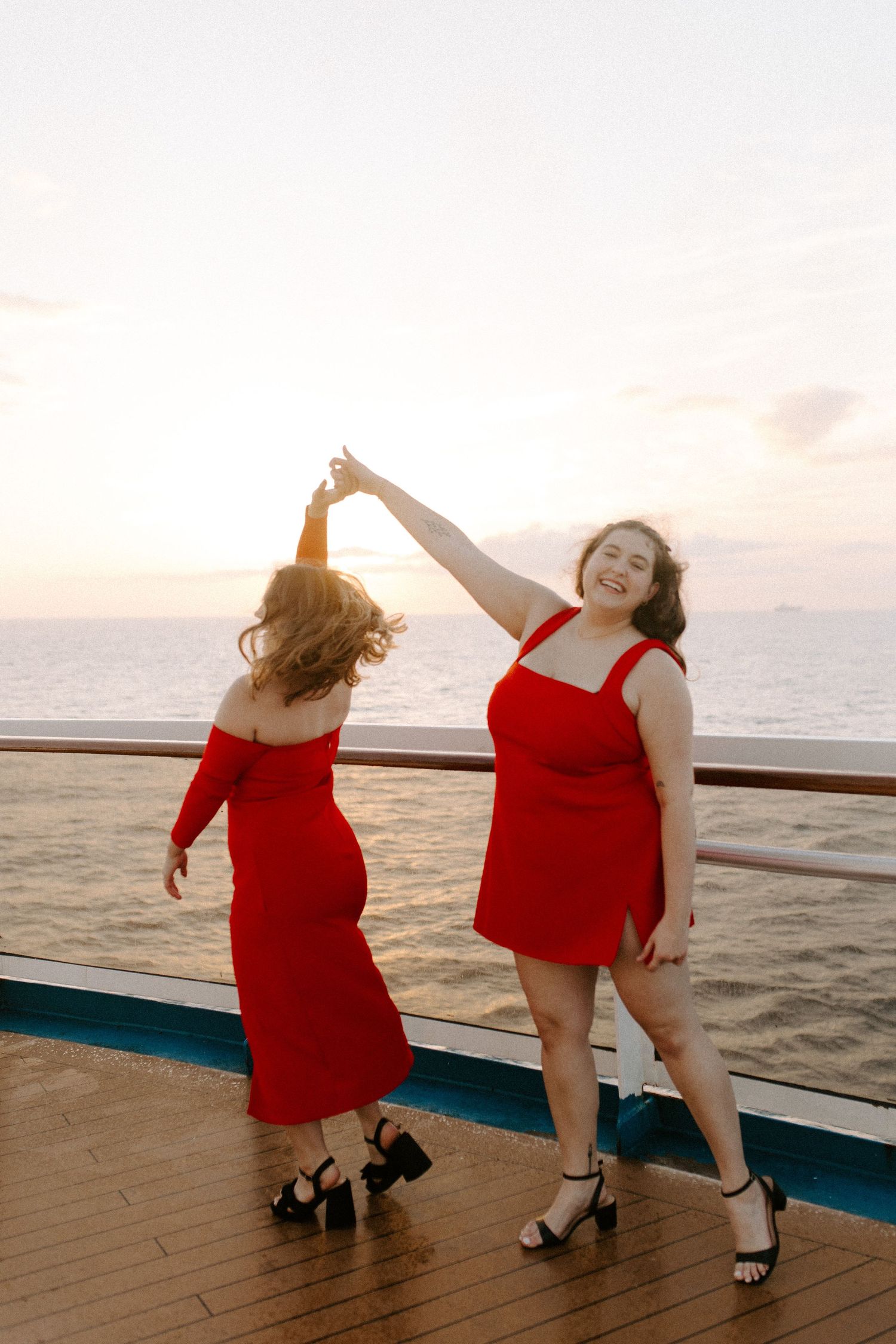 Friends twirling and dancing in red dresses on a cruise ship deck at dusk.