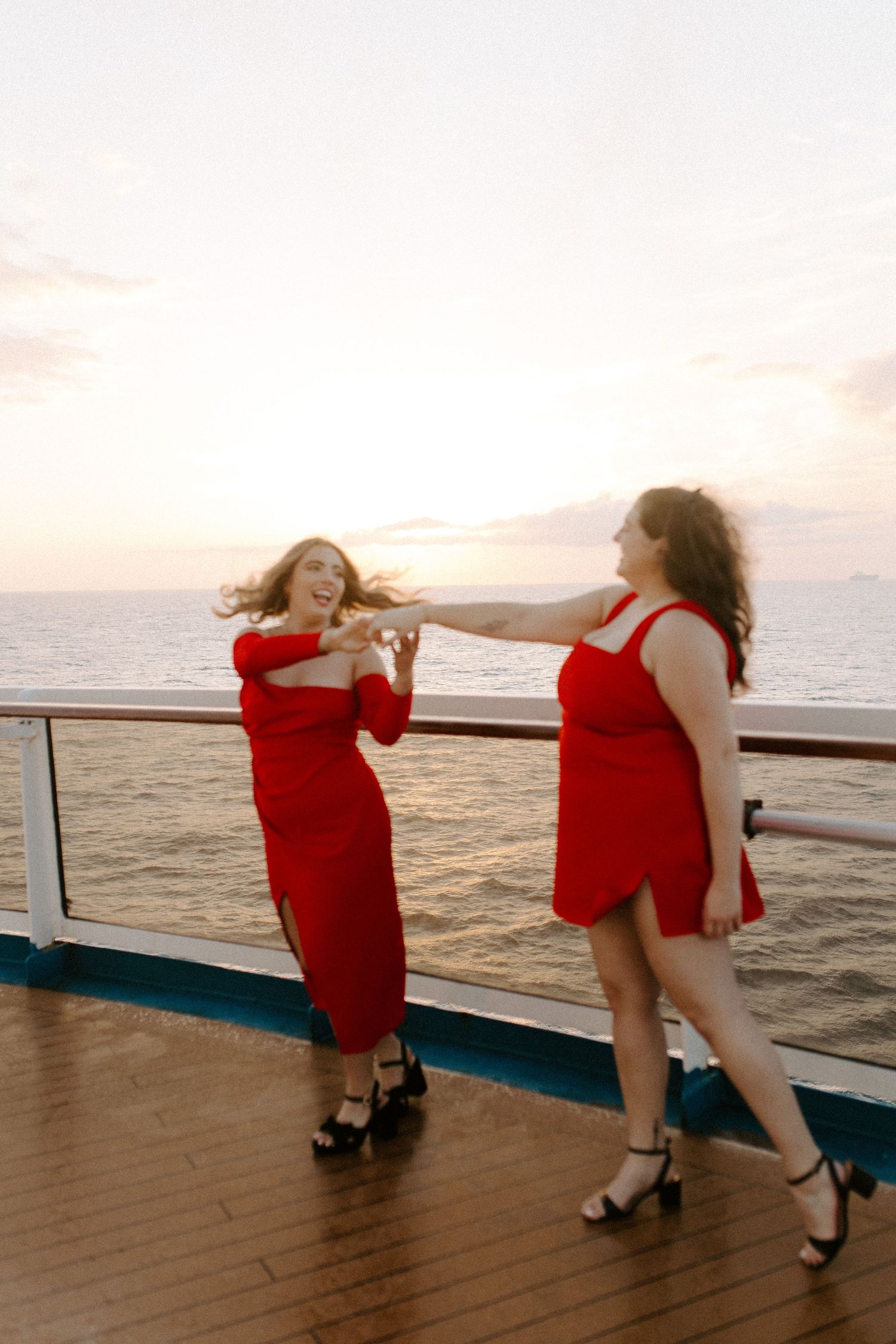 Two people in red dresses dancing playfully on a cruise ship deck at sunset.