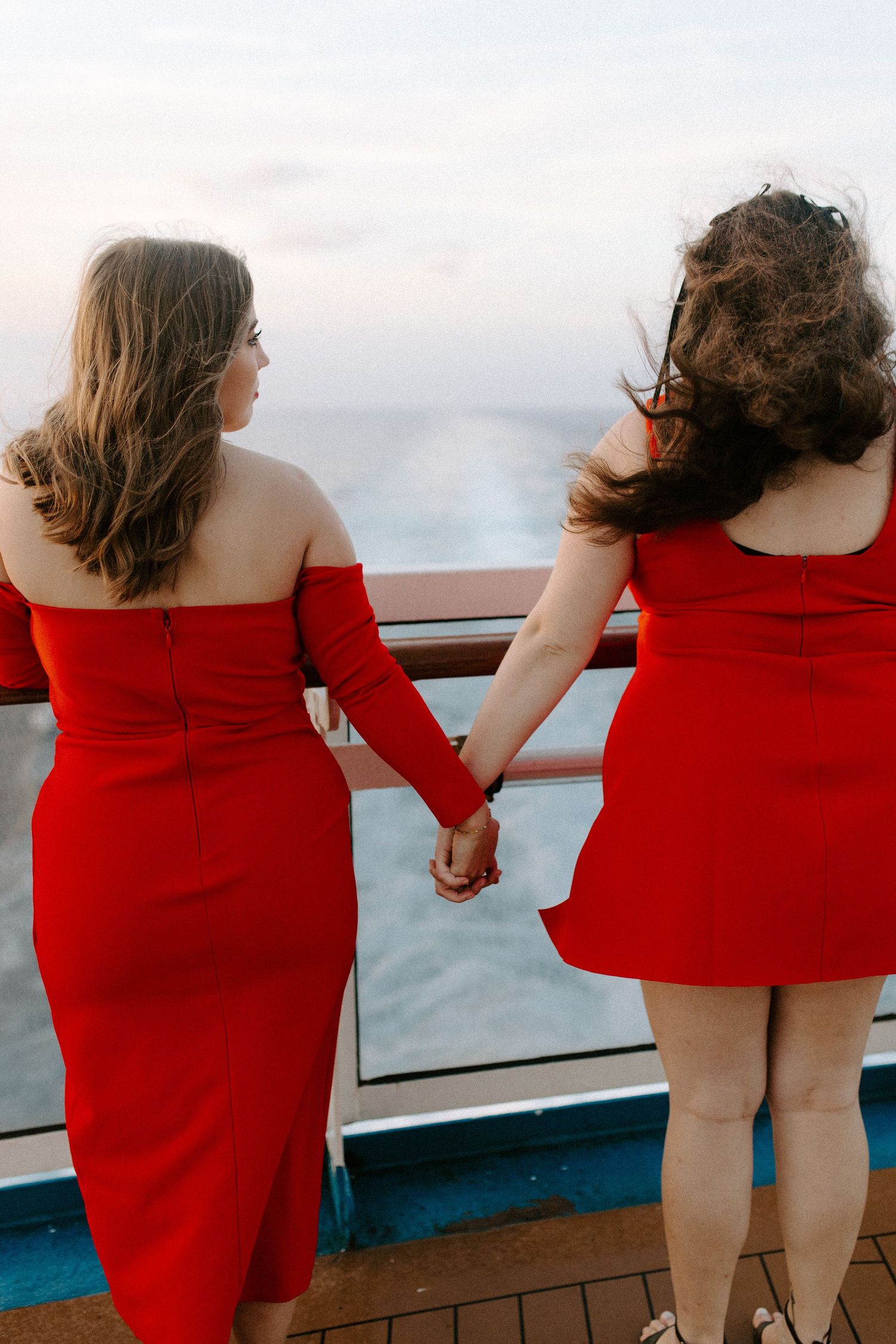 Two figures in matching red dresses hold hands while looking out over the ocean from a cruise ship deck at sunset.