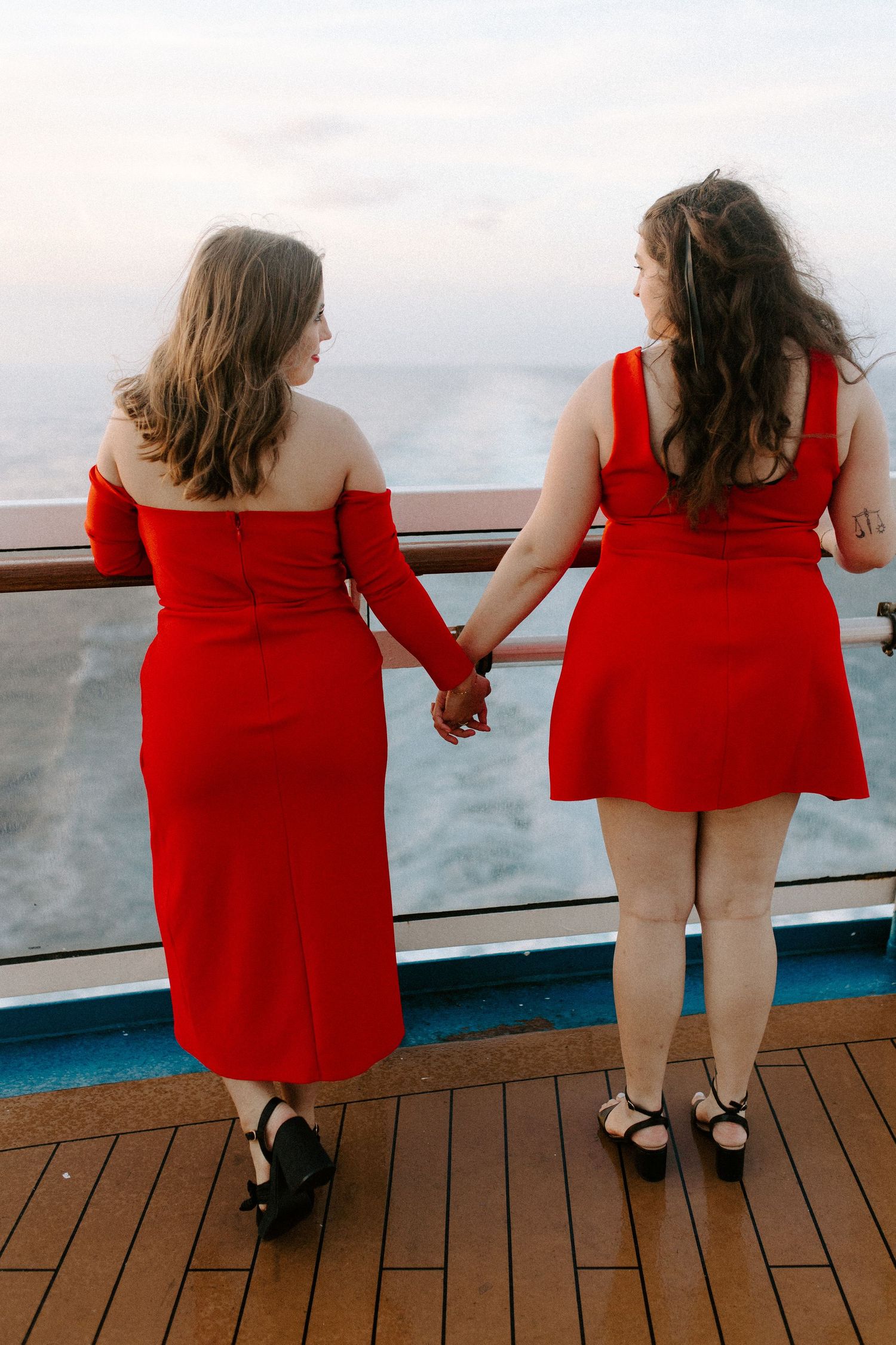 Two women in red dresses stand hand-in-hand on a wooden deck overlooking the ocean wake.
