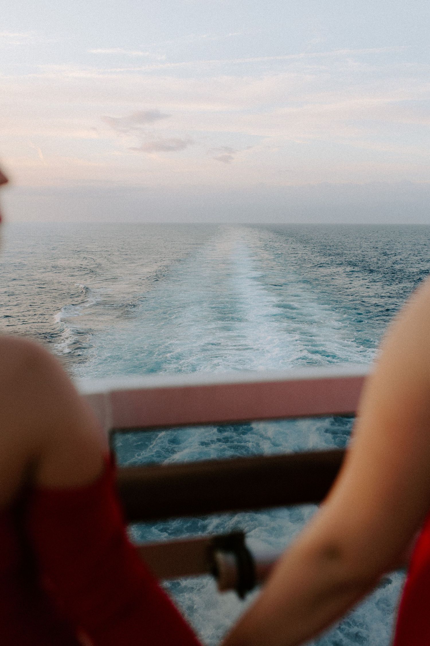 View of ocean wake and horizon from cruise ship deck with partial figures in red dresses visible.