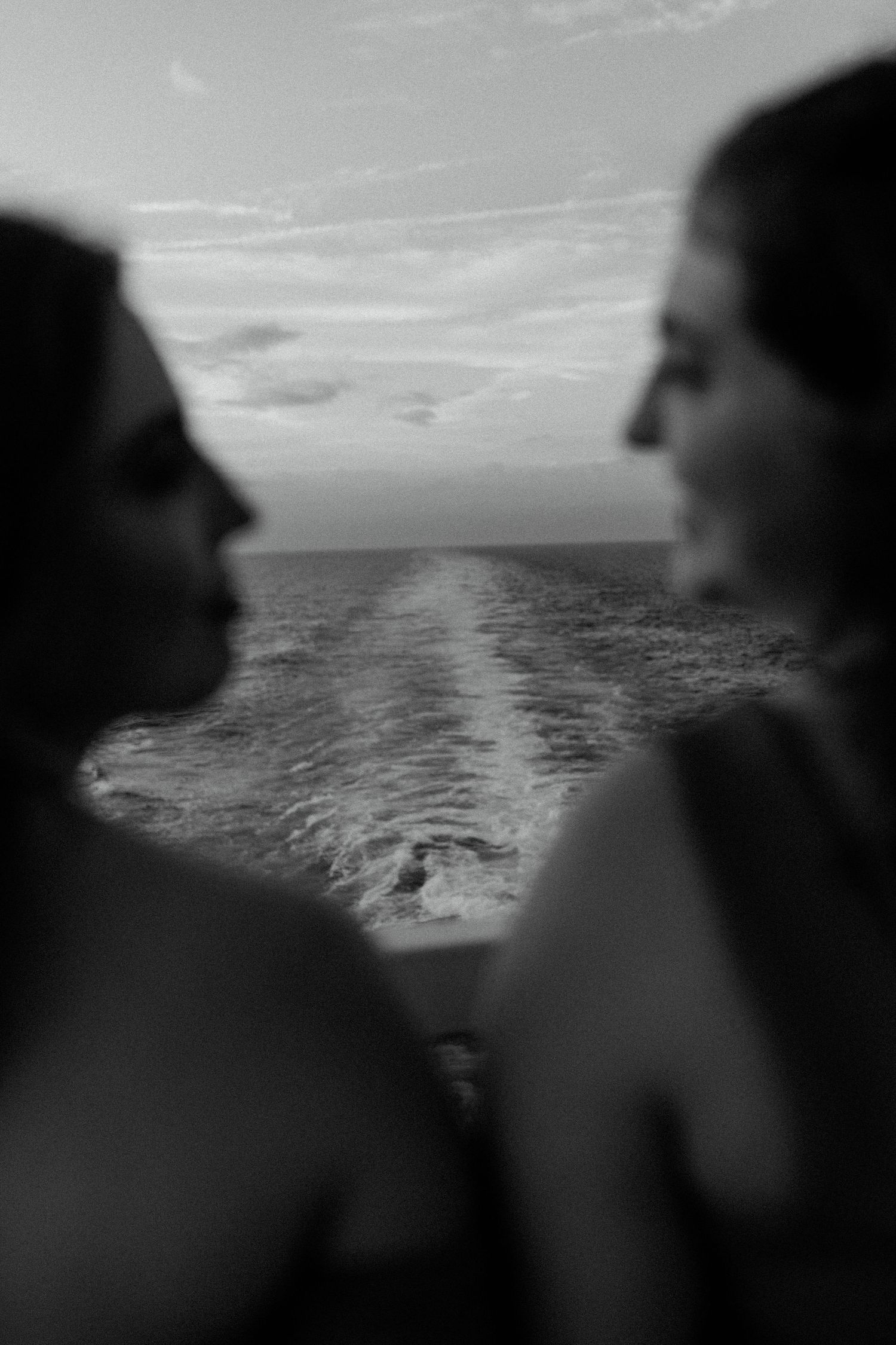 Black and white silhouette of two people looking at each other with ocean wake in background.