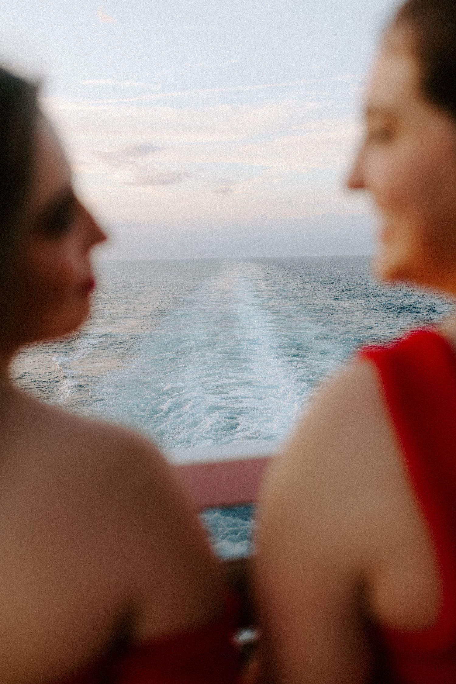 Two figures in red clothing look out over the ocean wake during sunset from ship's deck.