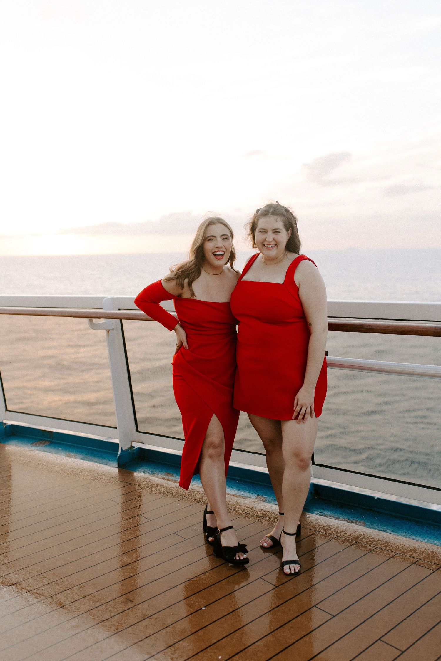 Two friends in matching red dresses pose together on cruise ship deck at sunset.