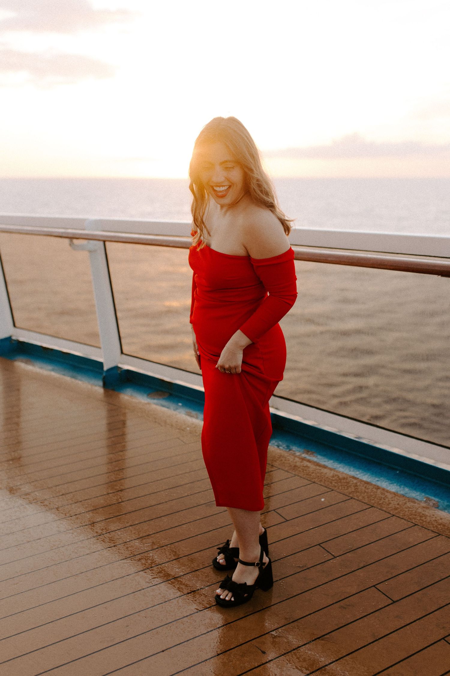Woman in red dress poses playfully against ship railing at golden hour.