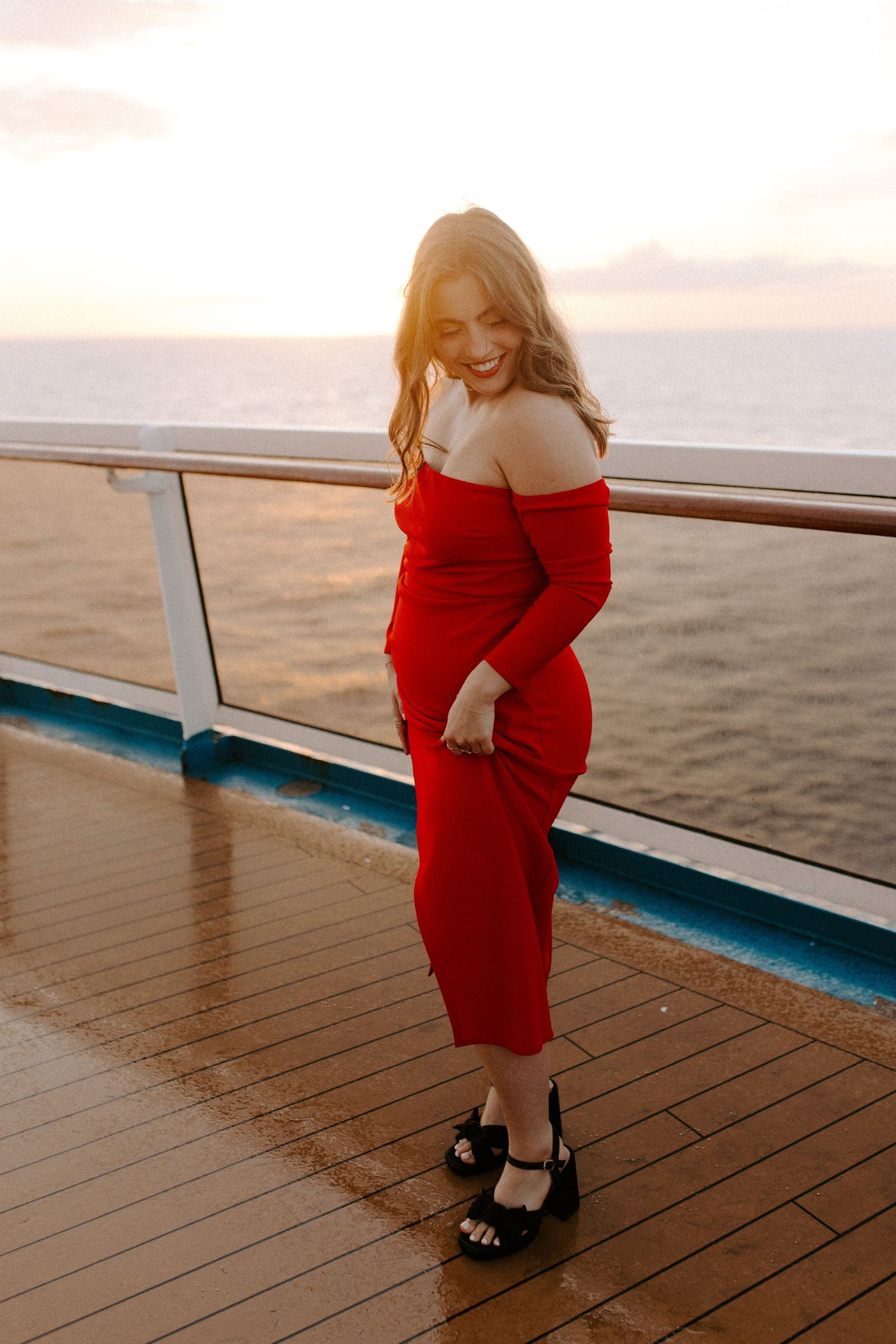 Woman wearing red off-shoulder dress smiling on wooden deck with ocean view.