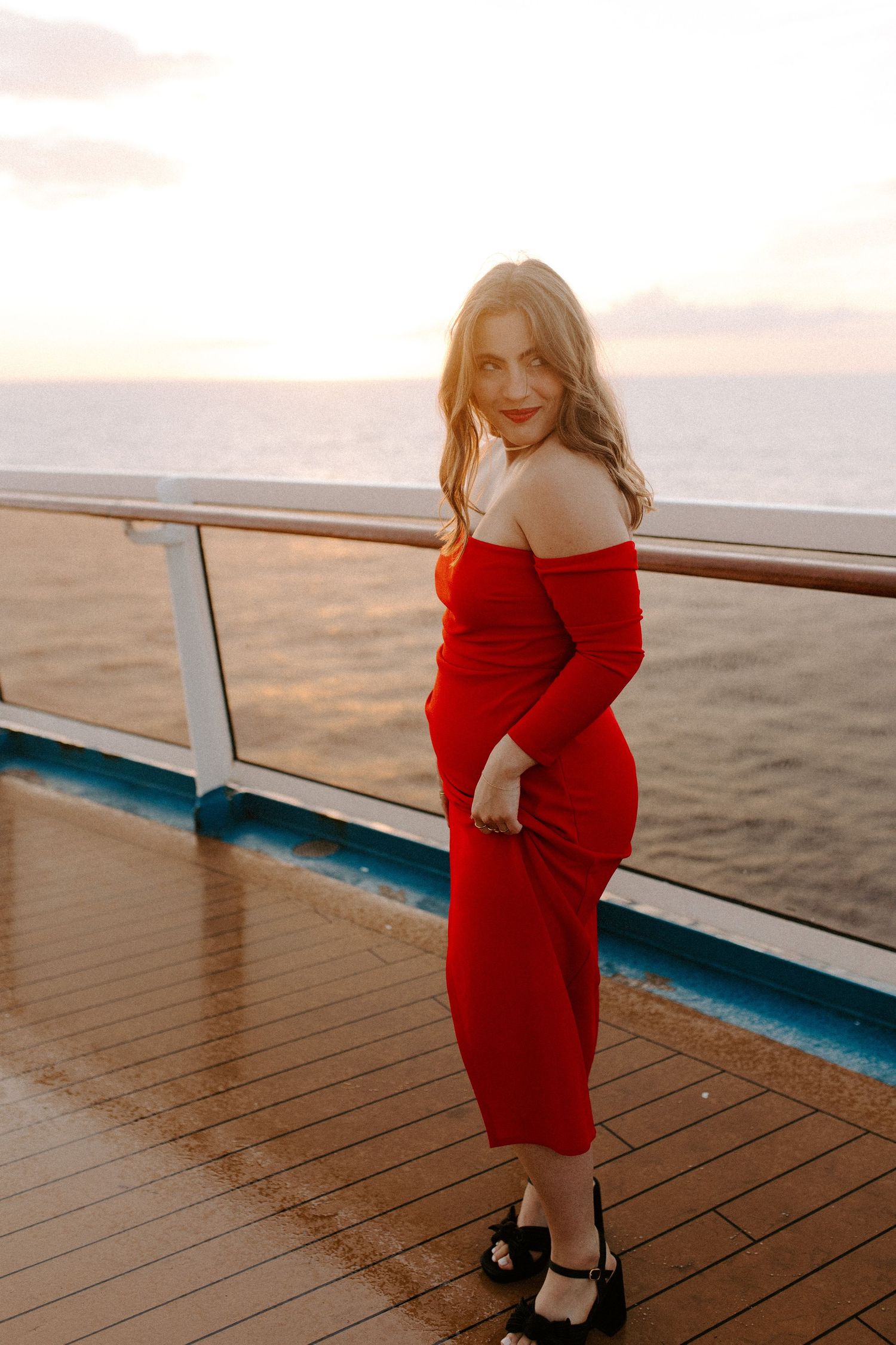 Woman in fitted red dress poses elegantly on cruise ship deck at dusk.