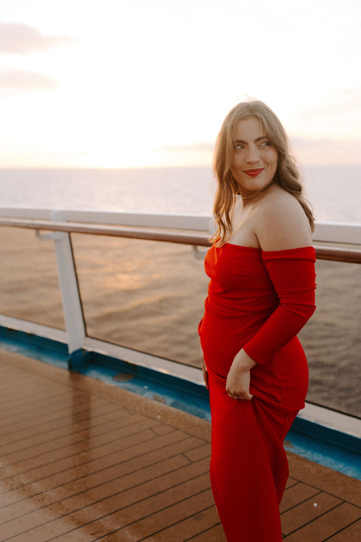 Woman in red evening dress stands against railing during romantic sunset cruise.