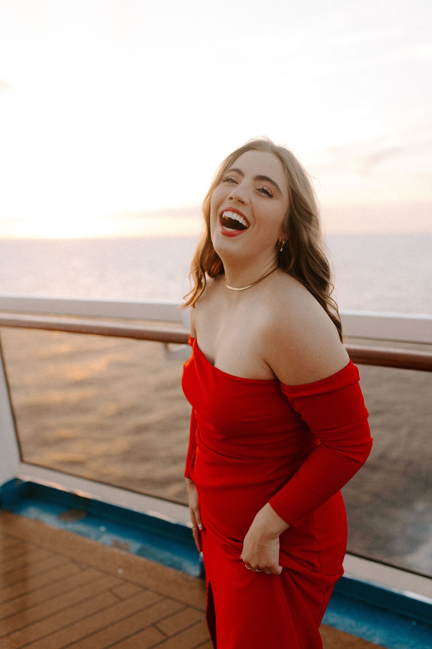 Woman in red dress laughing candidly during sunset photoshoot on ship deck.
