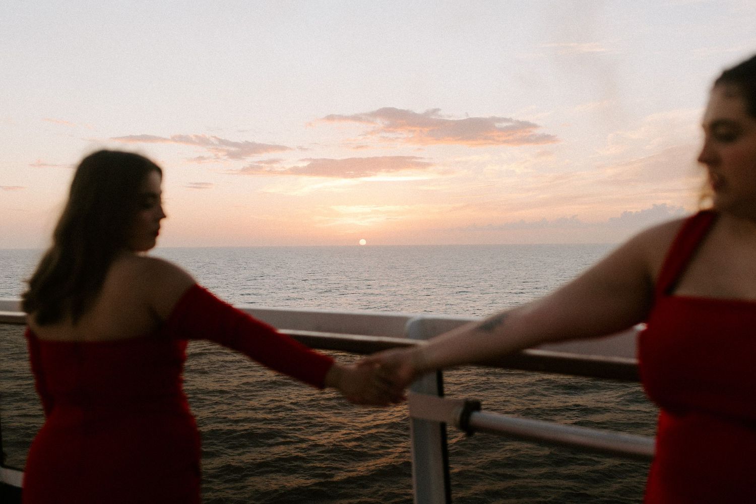 Two women in red dresses holding hands at sunset on cruise ship deck.