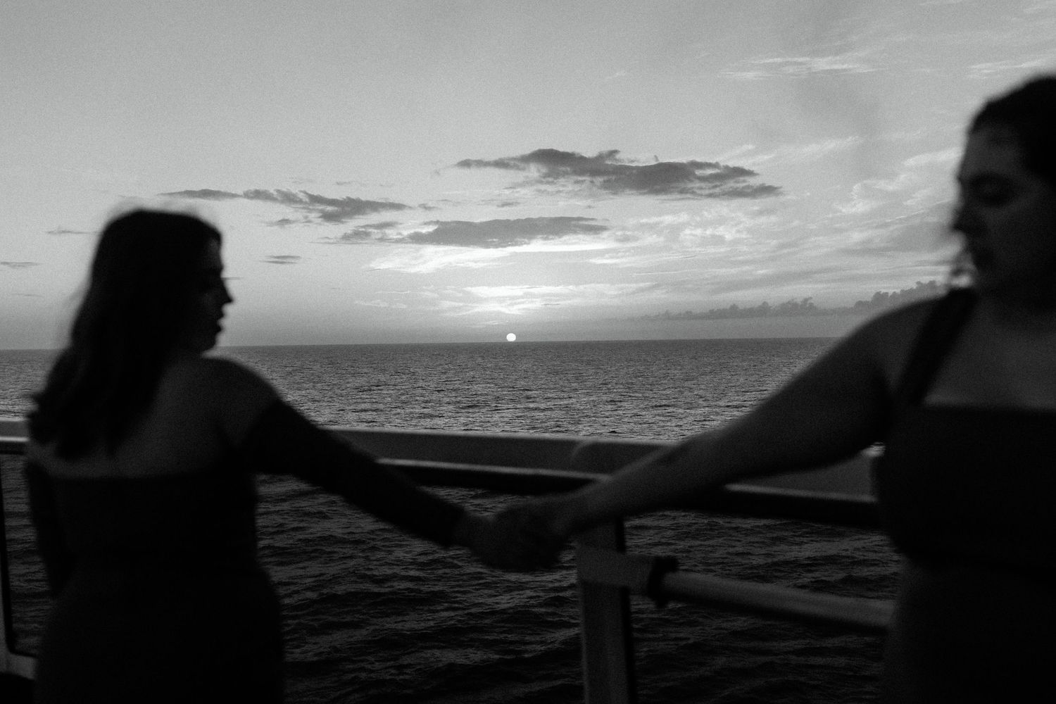 Silhouette of two women in dresses holding hands against sunset ocean view.