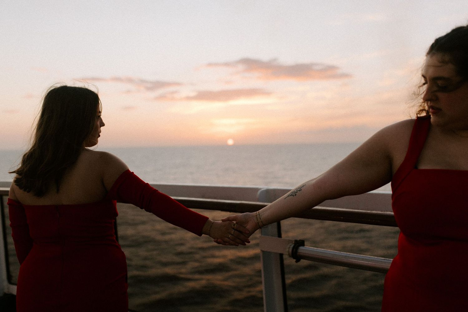 Women in red dresses hold hands while watching ocean sunset from ship deck.