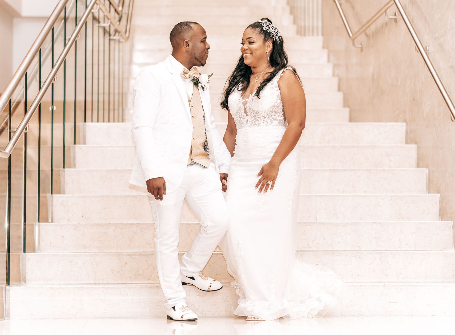 Wedding couple in all-white attire pose on elegant marble stairs.