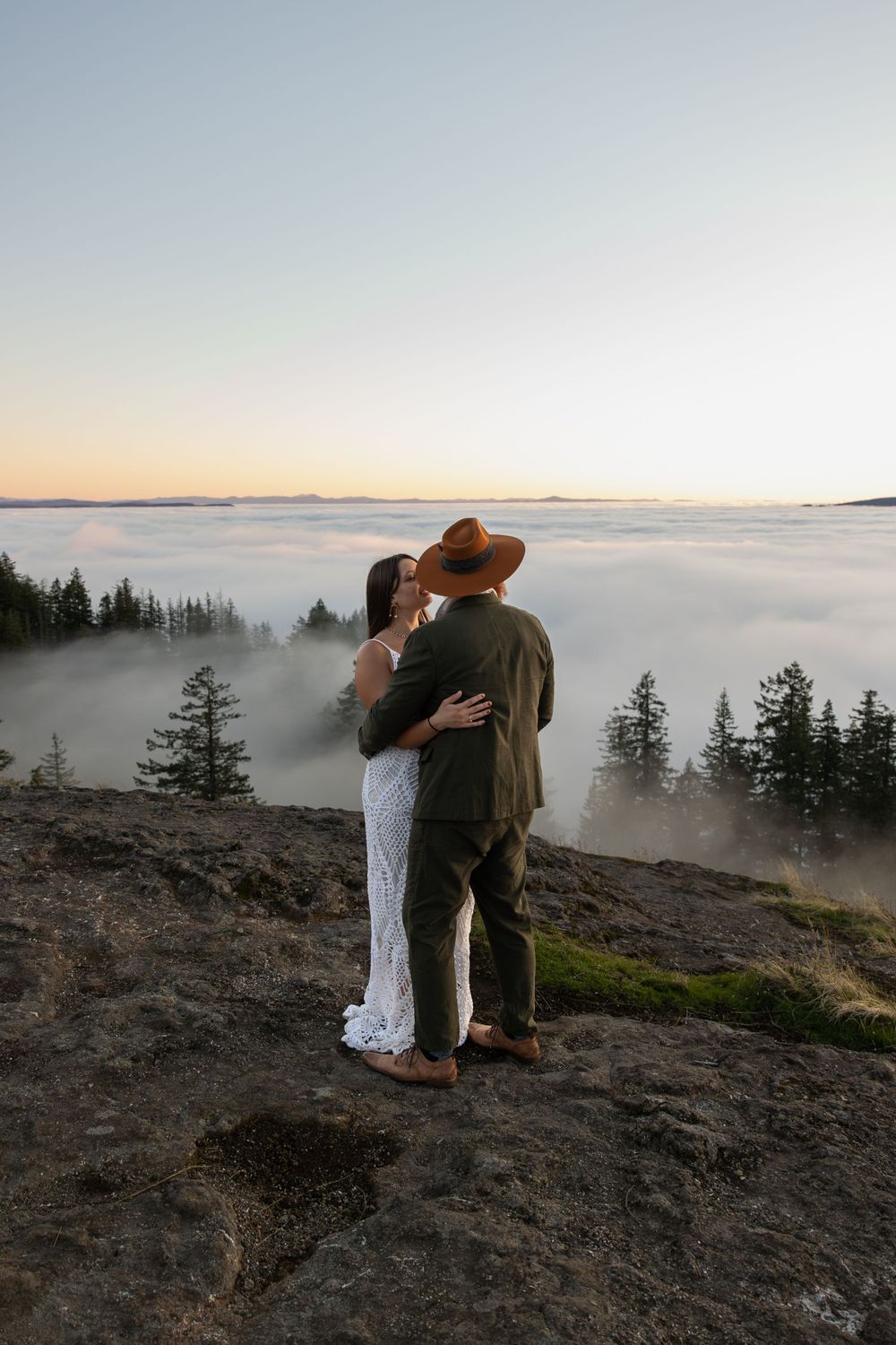 A couple embraces on a rocky cliff edge overlooking a misty valley at sunset.