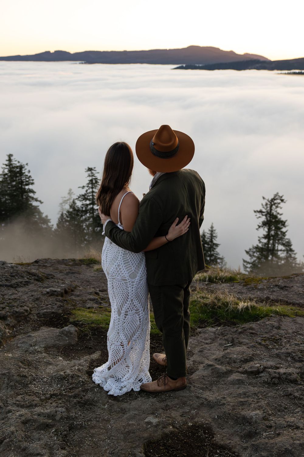 A couple stands together on a mountain edge overlooking a fog-filled valley at dusk.
