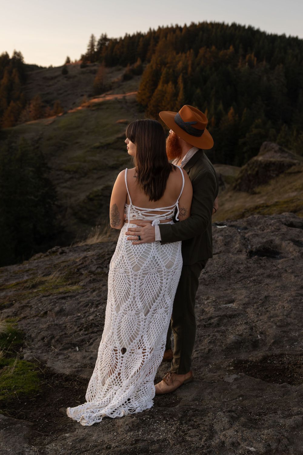 Couple embraces on mountainside at sunset while wearing bohemian wedding attire and holding vintage lanterns.