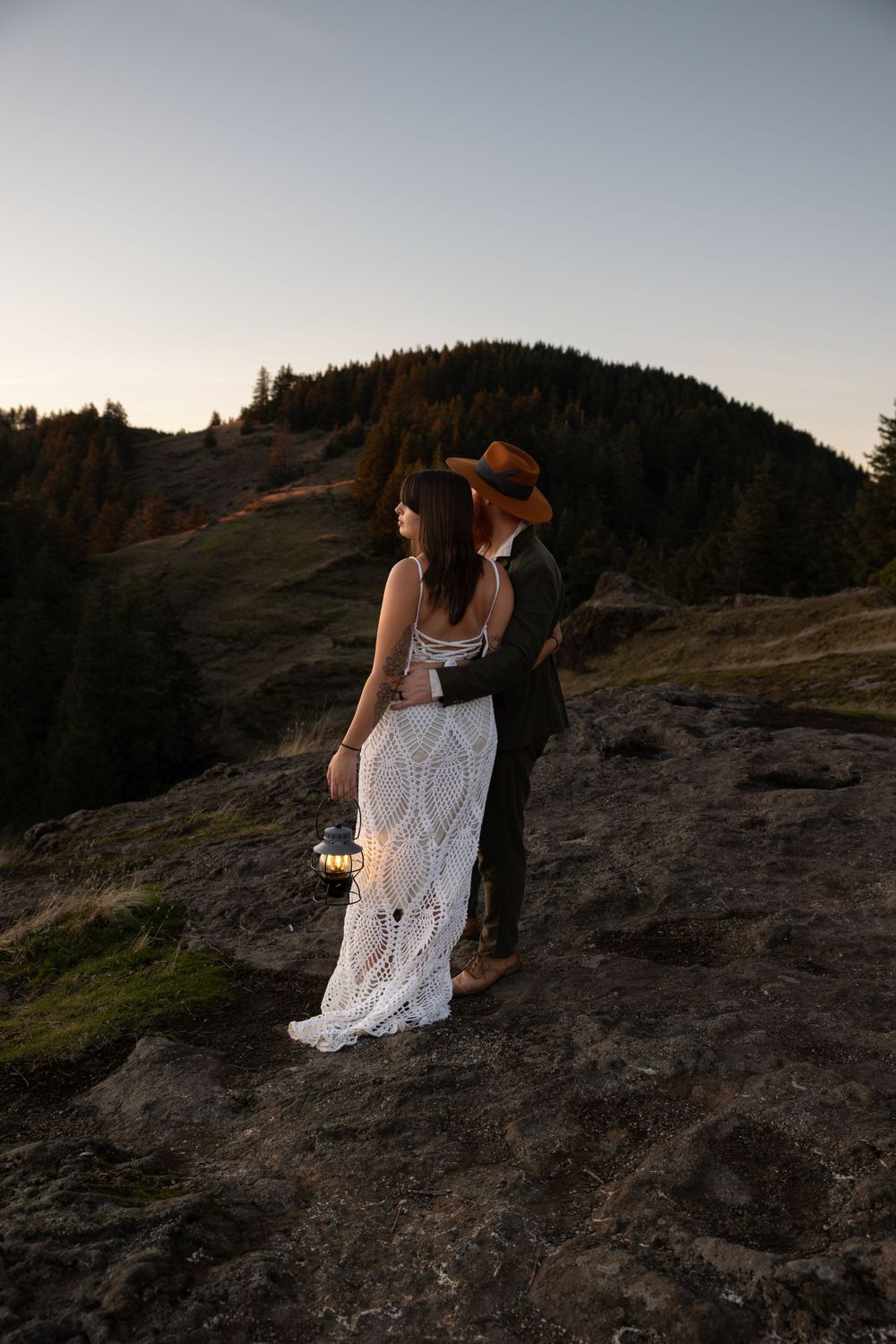 Romantic couple stands on rocky cliff edge at dusk in wedding attire with mountains in background.