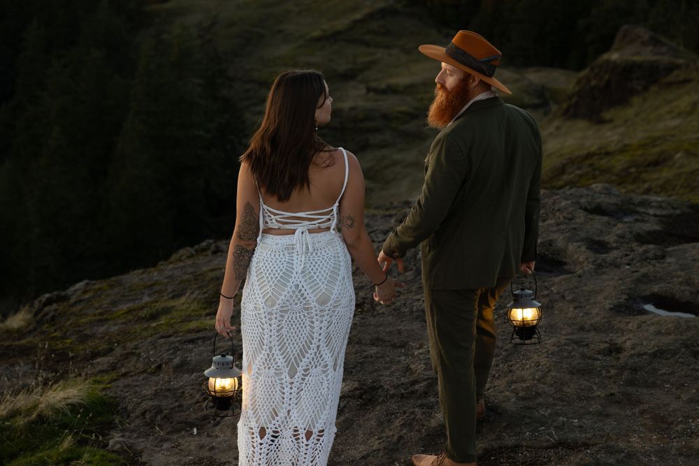 Couple in wedding attire holds lanterns while standing on mountain trail at twilight.