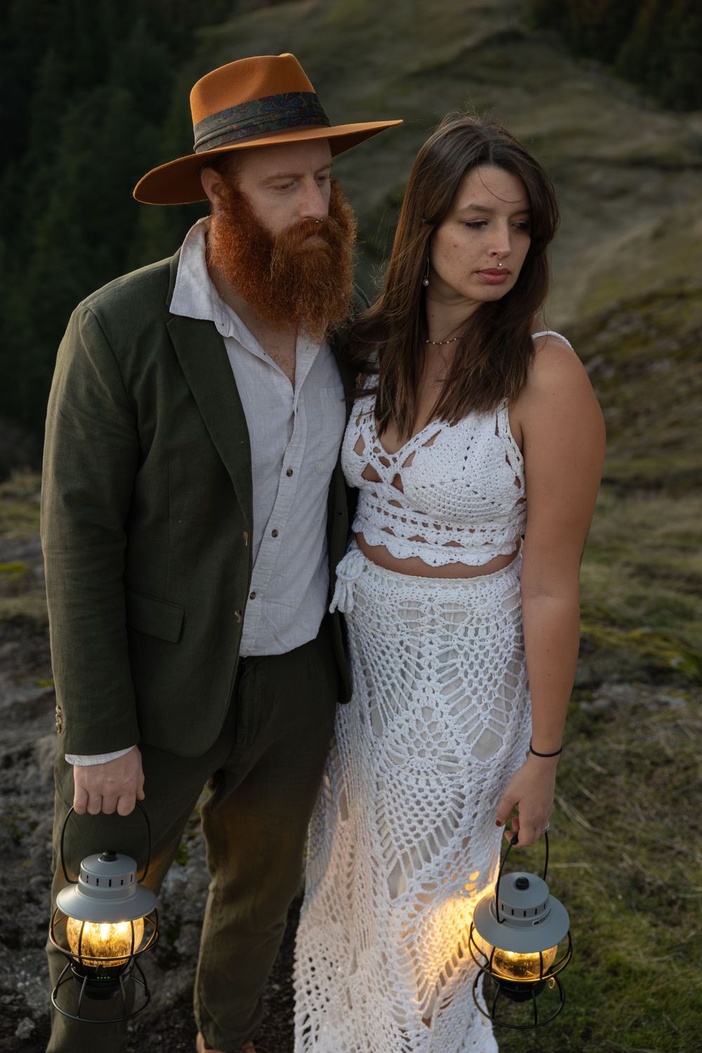 Couple in vintage-inspired wedding attire poses with lanterns on rocky mountain overlook at dusk.