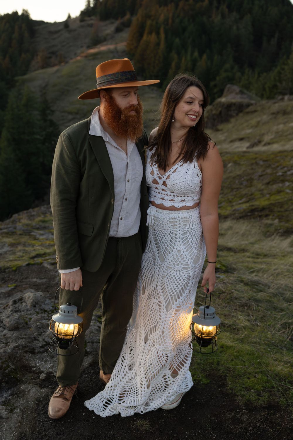 Couple stands together holding glowing lanterns on mountain cliff during sunset photoshoot.