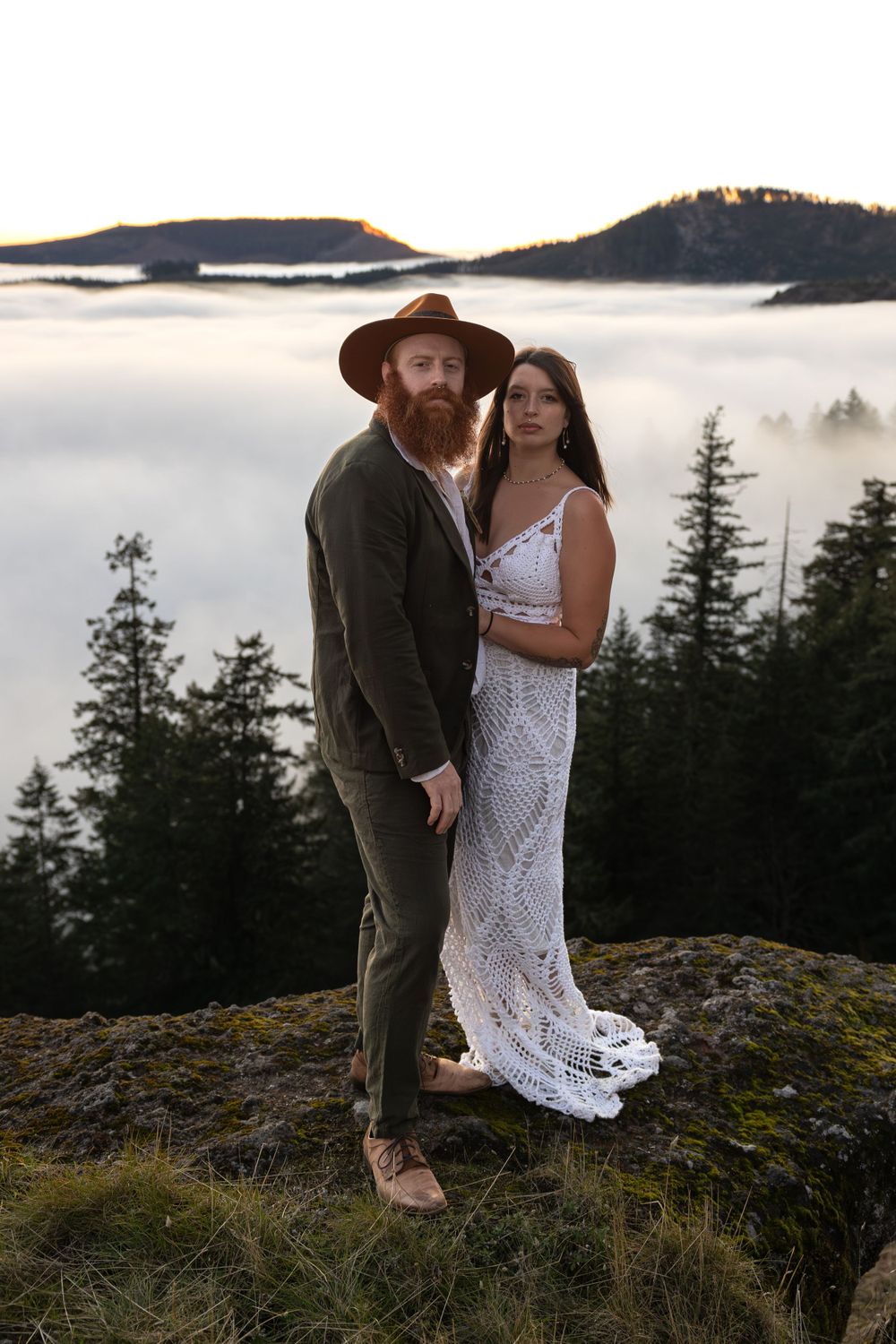 Couple poses on mountain peak above sea of clouds at sunset with vintage lanterns.
