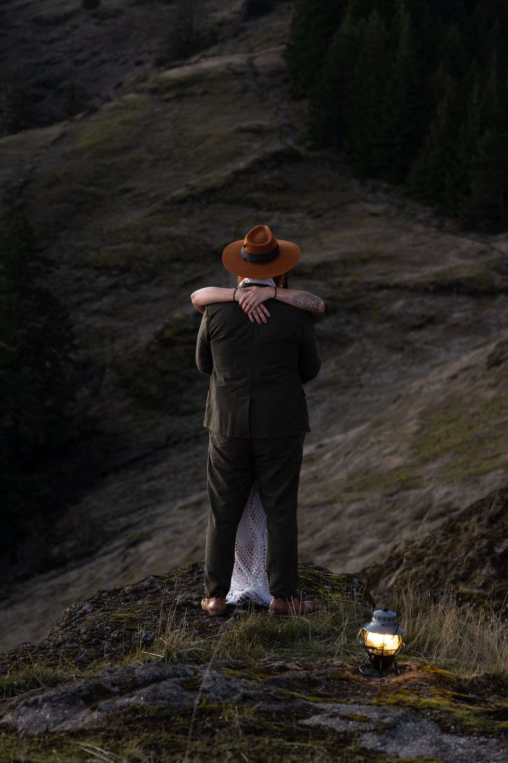 Figure in dark coat and brown hat embraces partner in white dress on mountainside with lantern below.