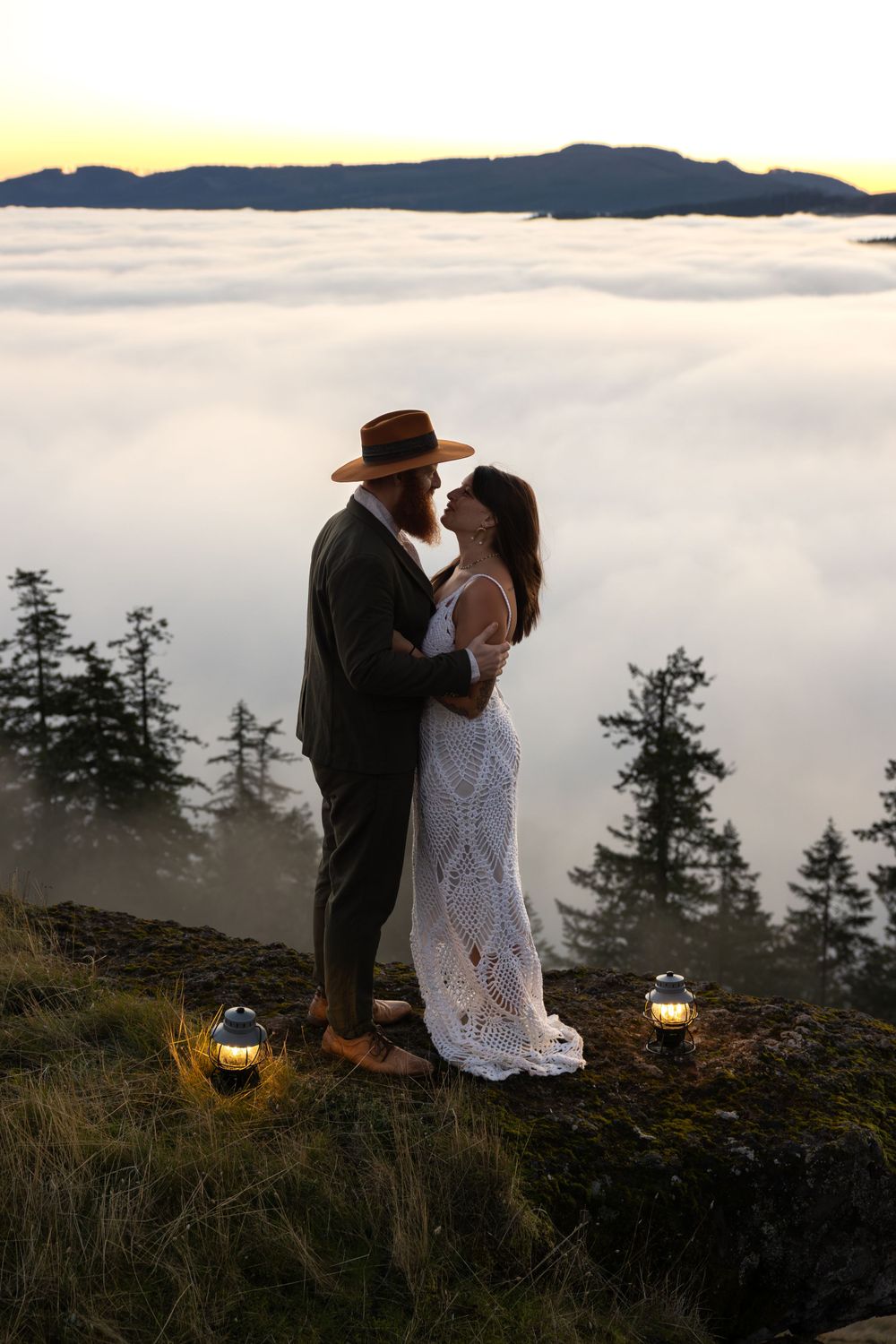 Couple shares intimate moment on mountain overlook above fog bank with lanterns glowing at sunset.