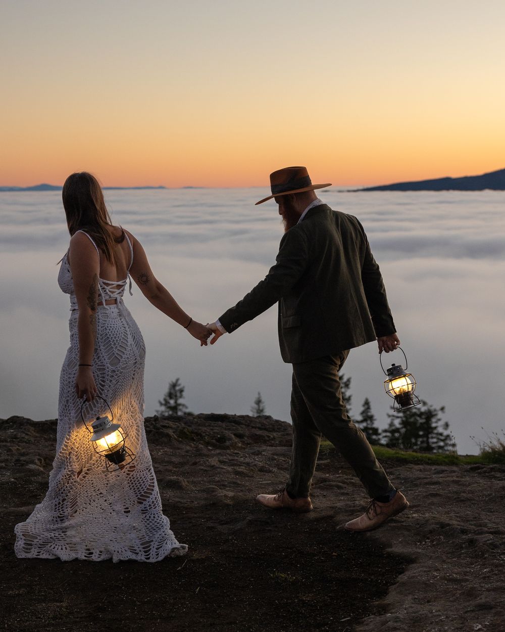 Couple holding lanterns stands on cliff edge above misty valley at sunset.