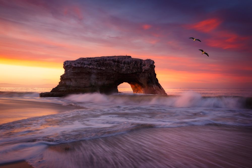 Natural stone arch formation at Natural Bridges State Beach in Santa Cruz during dramatic orange sunset.