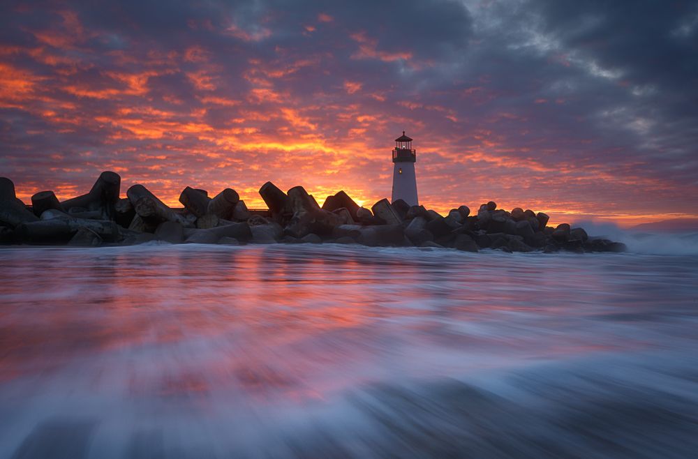 Lighthouse silhouetted against dramatic orange and purple sunset sky with reflective waters in the foreground.