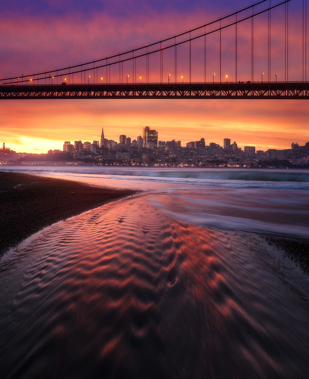 Dramatic sunset silhouettes San Francisco skyline under Golden Gate Bridge with rippled water reflection in foreground.