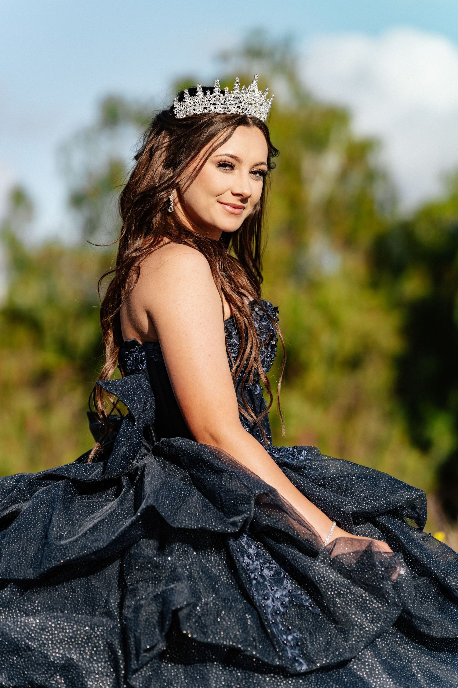 A person in a sparkly navy blue formal gown poses gracefully outdoors with a crown and flowing dress.