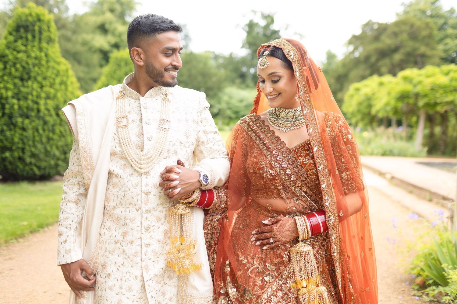 A couple in traditional South Asian wedding attire walks together in a garden setting wearing ornate cream and orange outfits.