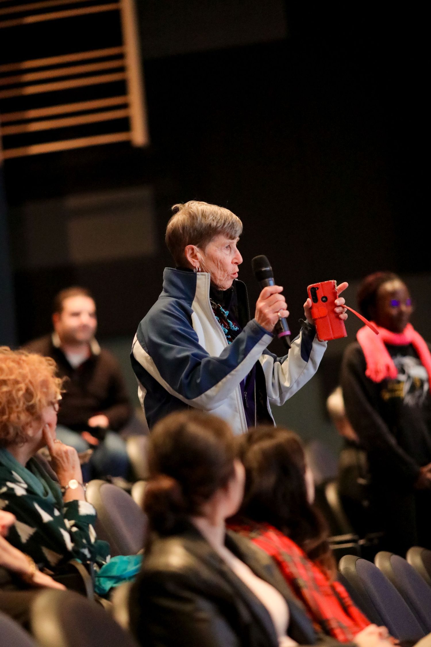 A person speaking with a microphone stands in an auditorium audience wearing a blue jacket holding a red puppet.