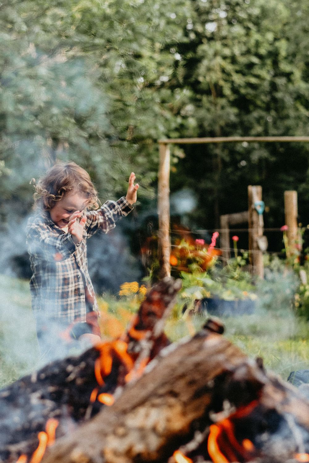 Person in plaid shirt dancing joyfully near a burning bonfire in a garden setting during daytime.