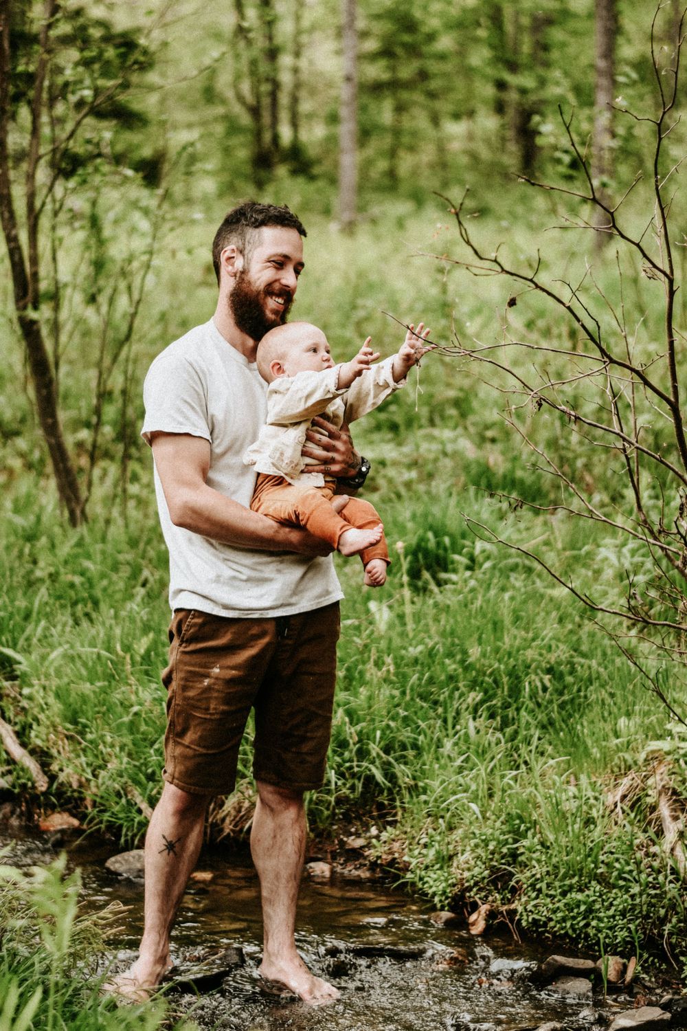 A parent and baby explore nature together while standing barefoot in a forest stream.