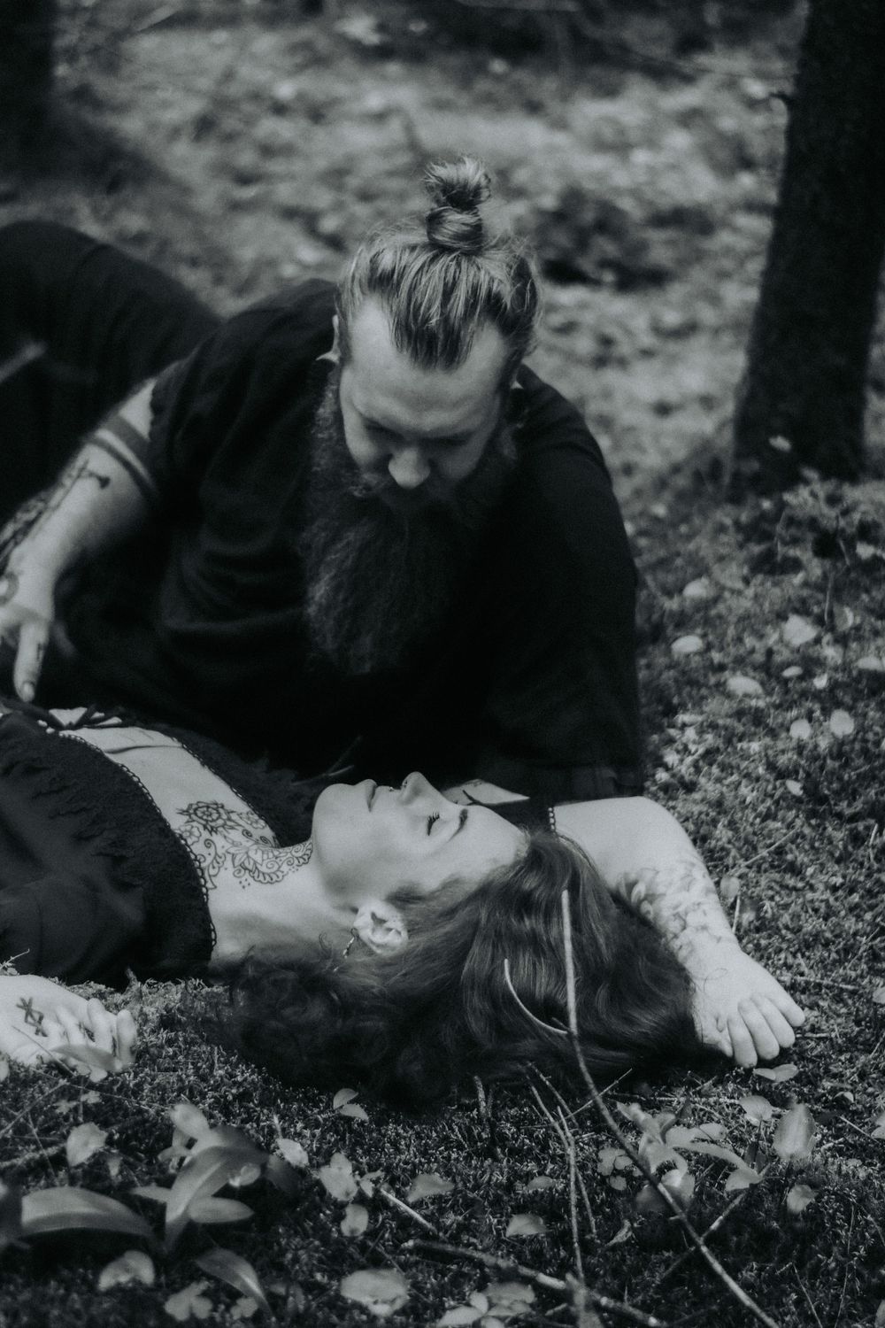 A black and white photograph showing two people in an intimate scene outdoors on the ground among leaves and grass.