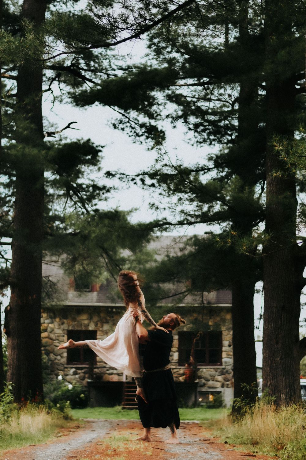 Two dancers perform an intimate lift on a forest path with a stone building in the background at dusk.